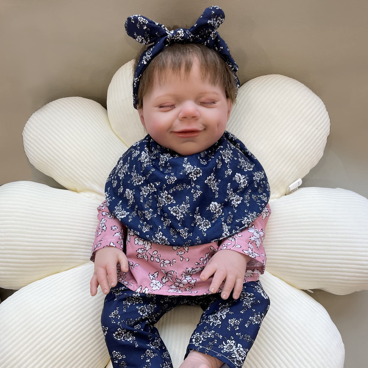 18" lifelike reborn baby doll with floral outfit and headband, smiling on plush flower cushion. Original product packaging included.
