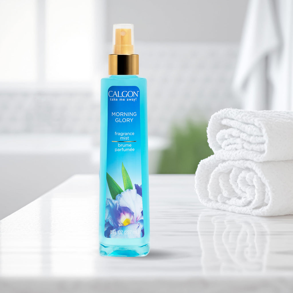 Calgon Morning Glory fragrance mist in a sleek blue bottle on a marble counter, accompanied by fluffy white towels for a refreshing experience.