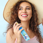 A smiling woman with curly hair and a straw hat holding Calgon Fragrance Body Mist, Morning Glor in original packaging.