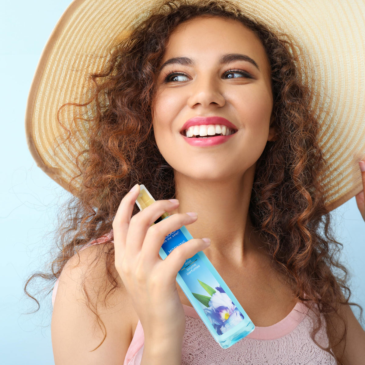 A smiling woman with curly hair and a straw hat holding Calgon Fragrance Body Mist, Morning Glor in original packaging.
