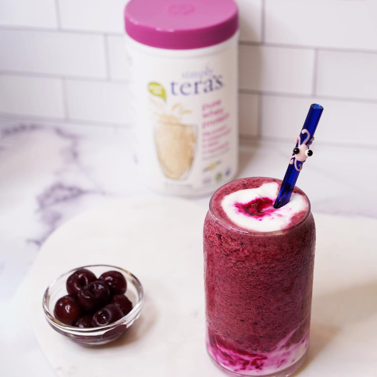 A refreshing berry smoothie topped with cream, next to Tera's Pure Whey Protein Powder packaging and a bowl of cherries.