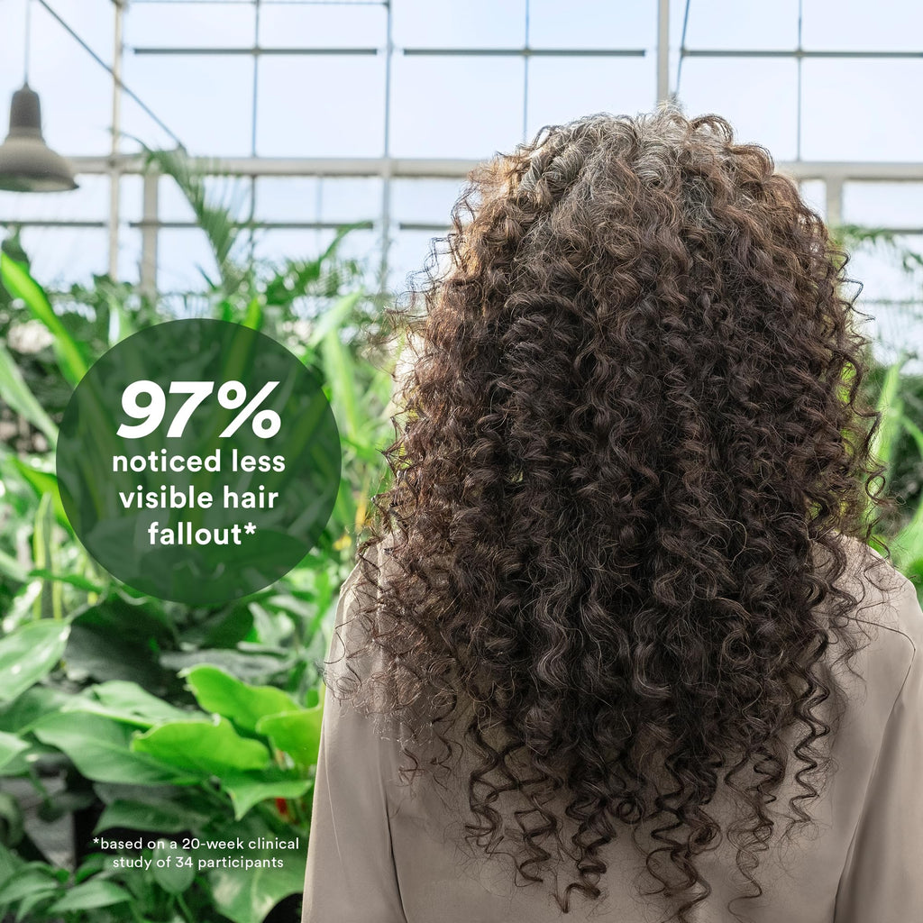 A person with curly hair stands amid green plants, highlighting the effectiveness of VERB Density Peptide Shampoo for reducing hair fallout.